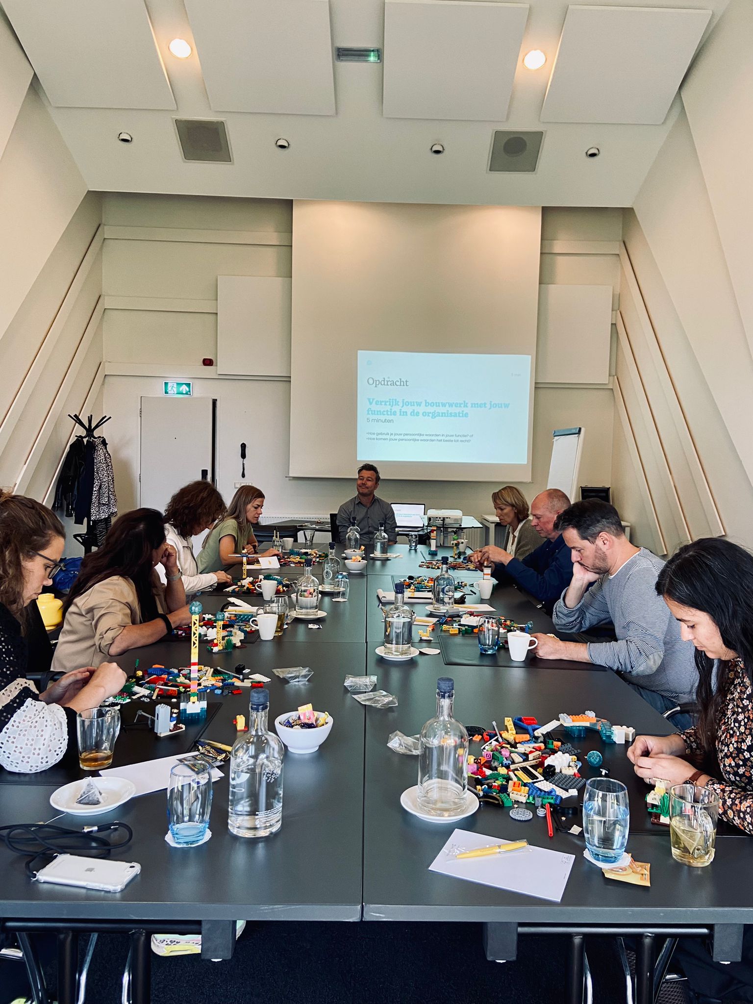 een groep mensen die aan een tafel zit met een persoon die vooraan aan de tafel staat